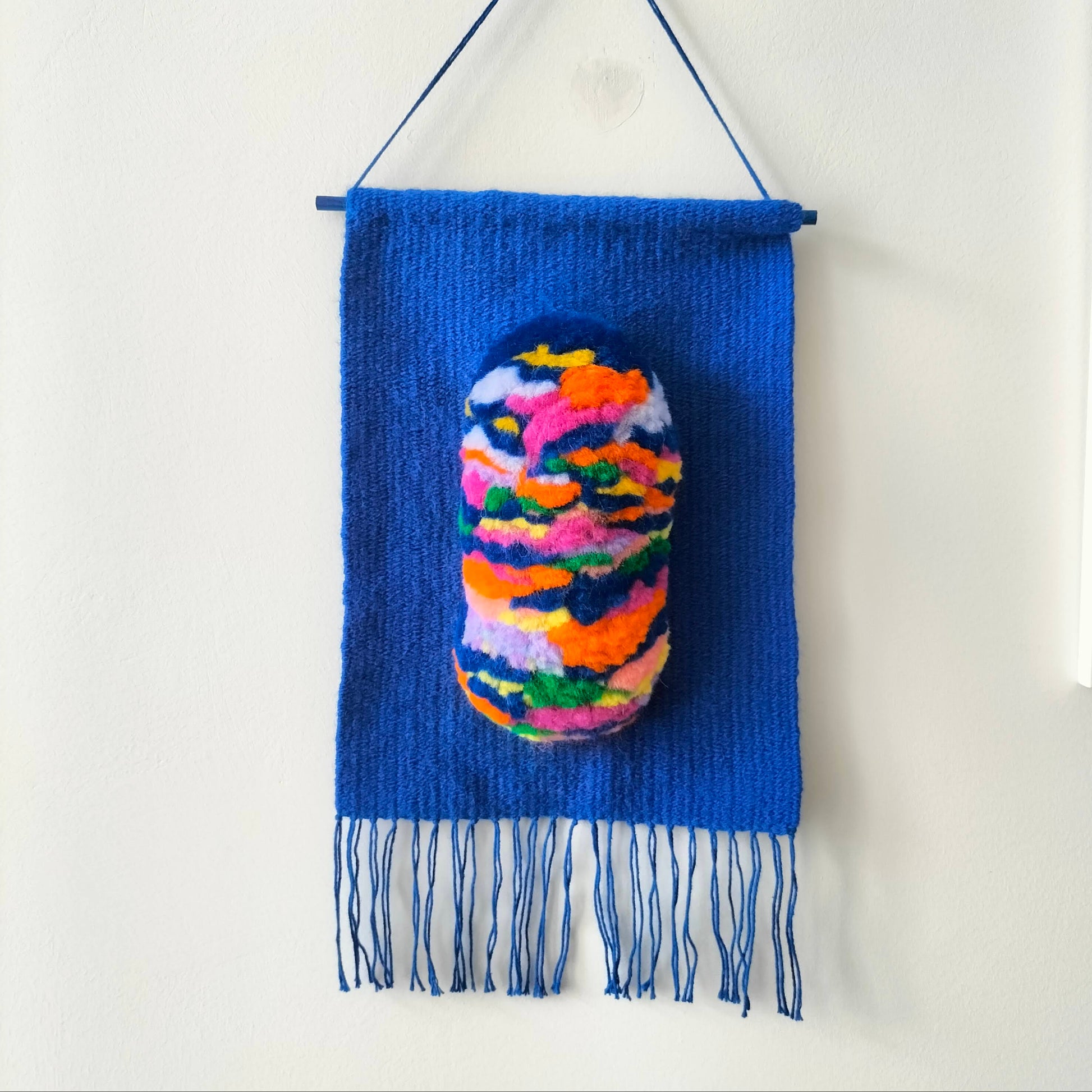 Blue woven wall hanging with colorful egg-shaped object on a white background